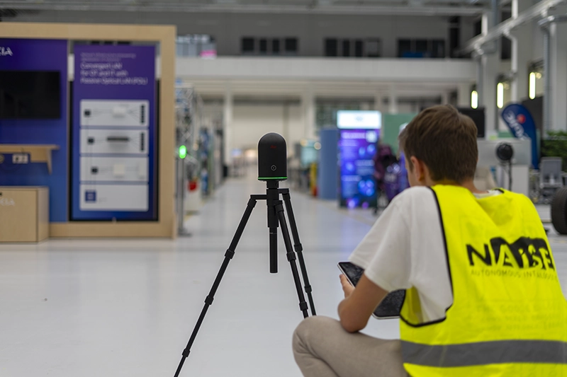 Technician in a NAiSE safety vest operating a tripod-mounted 3D scanning device in a large industrial hall to capture the facility for digital twin mapping.